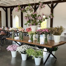 Load image into Gallery viewer, Decorative floral arrangements on a long wooden table in a room with high ceilings and chandeliers.
